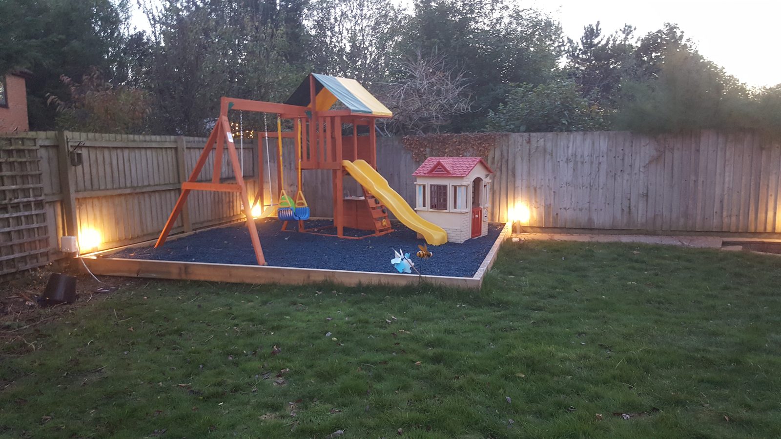 Outdoor garden lighting illuminating a family play area and surrounding garden at dusk.